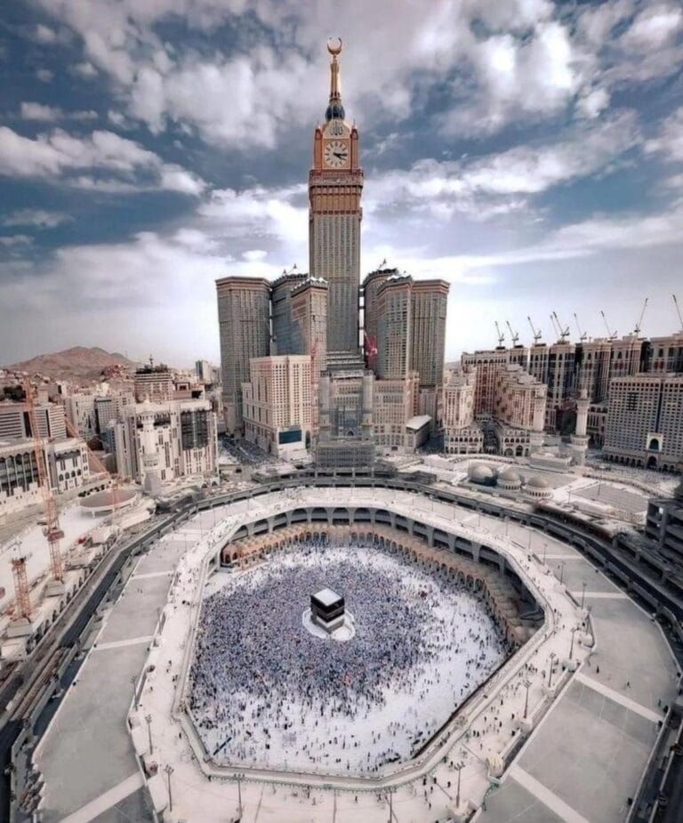 Aerial view of the Masjid al-Haram in Mecca showing thousands of pilgrims circumambulating the black Kaaba in the central courtyard, with the tall Abraj Al-Bait Clock Tower and surrounding cityscape under a partly cloudy sky.