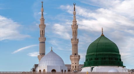 Mosque skyline featuring a prominent green dome next to a white dome and two ornate twin minarets set against a partly cloudy blue sky.