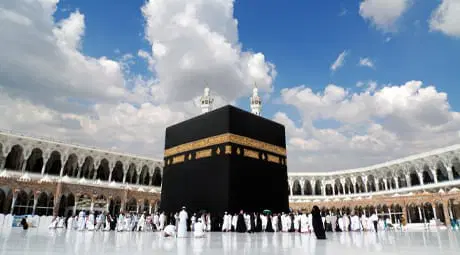 Black-clad Kaaba with a gold-embroidered band at the center of the white-marble courtyard of the Grand Mosque, surrounded by pilgrims and encircled by arcaded colonnades beneath a blue sky with scattered clouds.
