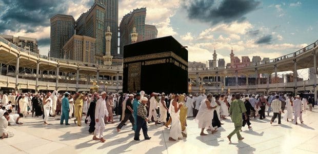 Crowds of pilgrims walk around the black, gold-embroidered Kaaba in the open courtyard of Masjid al-Haram, framed by mosque colonnades and towering modern buildings under a dramatic cloudy sky.