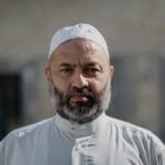 Middle-aged man with a salt-and-pepper beard and serious expression, wearing a white skullcap and light-gray buttoned shirt, facing the camera with a softly blurred stone background.