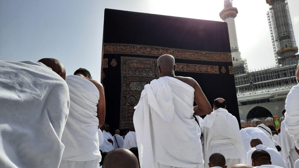 Pilgrims dressed in white ihram garments stand facing the Kaaba — the large black cube draped in a gold-embroidered kiswah — with mosque minarets and scaffolding visible in the background.