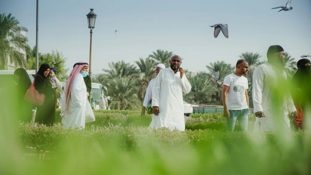 Group of people in a sunlit green park with palm trees and pigeons overhead, centered on a man in a white thobe touching his face while others — women in black abayas, a man in a red-and-white keffiyeh, and a casually dressed man — stand or walk nearby.