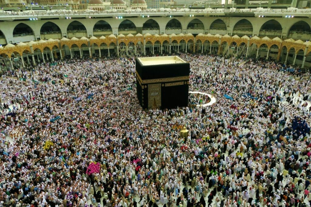 The Kaaba, a black cube draped in a gold-trimmed cloth, surrounded by thousands of pilgrims circumambulating in the open courtyard of Al-Masjid al-Haram with arched colonnades and balconies in the background.