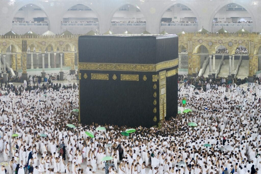 Pilgrims in white ihram circumambulating the black, gold-embroidered Kaaba at the Grand Mosque in Mecca, many holding green umbrellas in light rain beneath ornate arches.