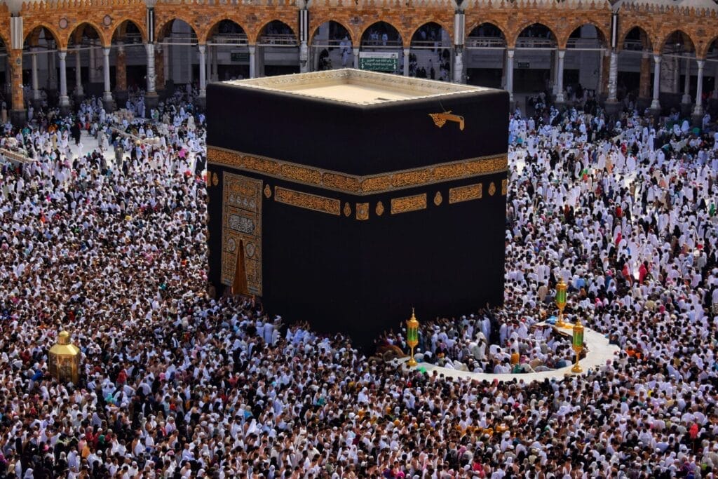 The Kaaba, a large black cube draped in a gold-embroidered cloth, surrounded by thousands of white-clad pilgrims performing tawaf in the courtyard of the Grand Mosque with arched colonnades visible in the background.