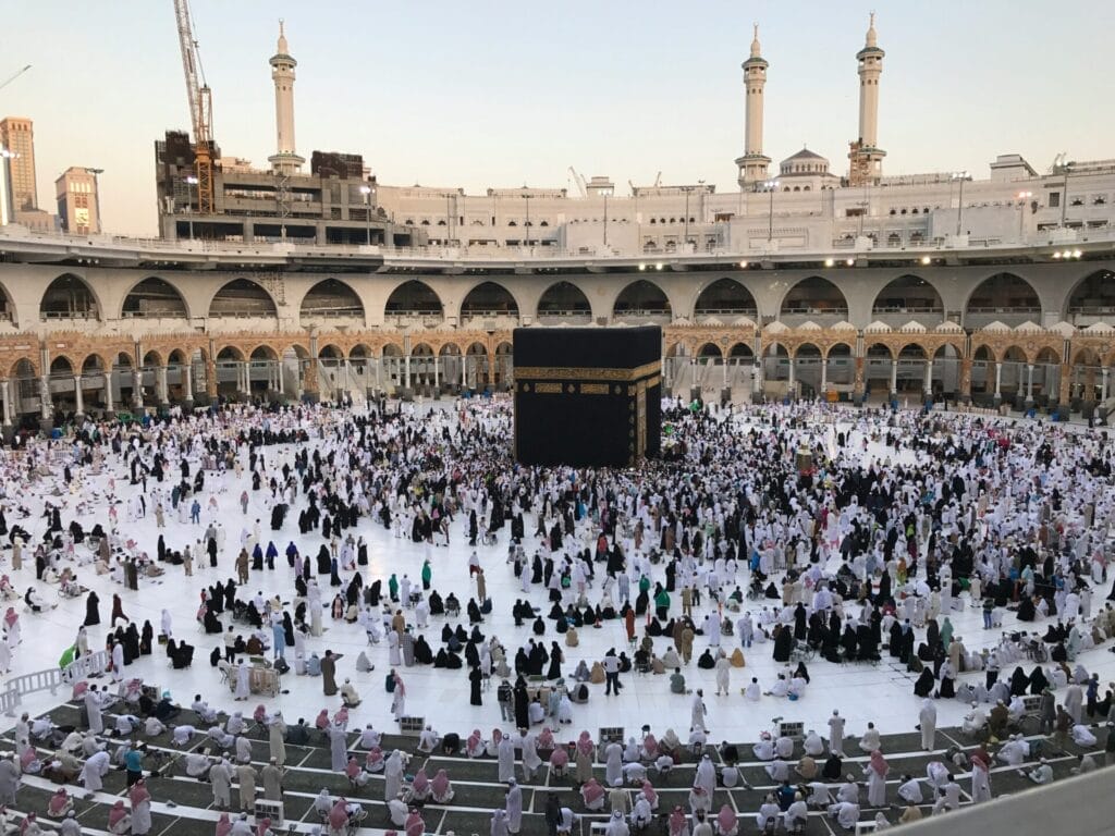 Aerial view of the Kaaba, the black cube draped in a gold-embroidered cloth, at the center of Masjid al-Haram's white-marble courtyard with thousands of pilgrims in white and dark robes circling it beneath surrounding arches and tall minarets.