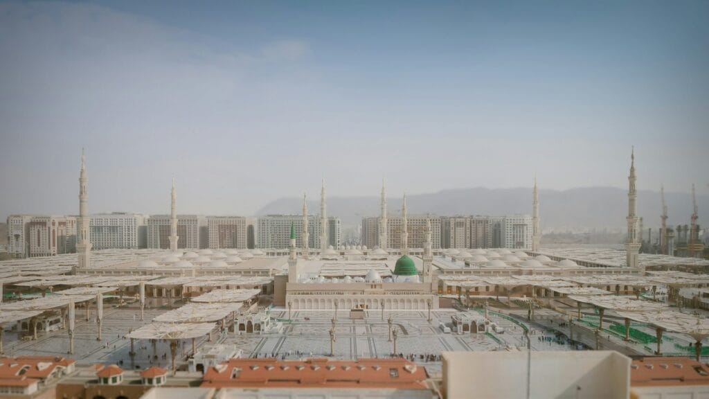 A wide aerial view of Al-Masjid an-Nabawi in Medina showing the mosque's central green dome, multiple tall minarets, rows of large retractable white umbrellas shading the courtyard, surrounding multi-story buildings and distant mountains under a pale blue sky.